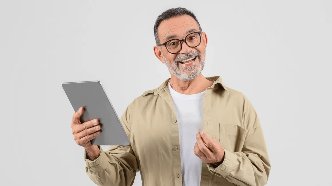 Mężczyzna w średnim wieku z brodą i okularami trzyma tablet i uśmiecha się do kamery, ubrany w beżową koszulę i białą koszulkę, na jasnym tle